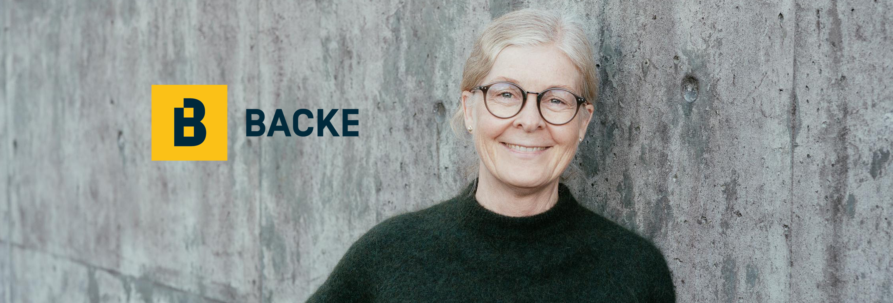 Smilende dame med briller foran grå murvegg