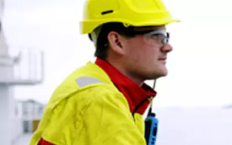 Mann i gul og rød kjeledress og gul hardhat som ser utover havet